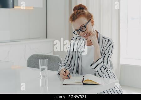 Europäische Frau macht Notizen in persönlichen Planer lächelt sanft hält Stift Posen auf dem Schreibtisch Stockfoto