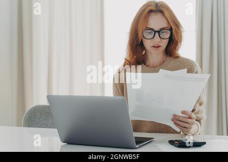 Serious Rotschopf kaukasischen Frau berechnet Rechnungen hält Papierdokumente hat fokussierten Blick Stockfoto