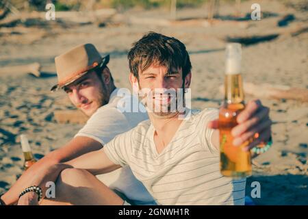 Portrait von Freunden, die sich am Strand ausruhen und Bier trinken. Fröhliche junge Leute, die eine schöne Zeit miteinander verbringen, während sie am Strand sitzen und lächeln Stockfoto