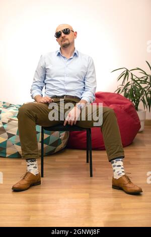 Stilvoller Mann mittleren Alters, der auf einem schwarzen Stuhl sitzt. Schwarze Brillen verleihen seinem Gesicht etwas Ernsthaftigkeit. Fotoshooting-Tag für Magazin in modernem Interieur Backgro Stockfoto