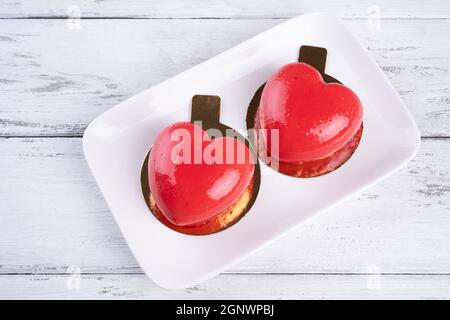 Zwei Mousse-Kuchen in Form eines roten Herzens auf einem weißen Teller auf weißem Holzgrund. Stockfoto