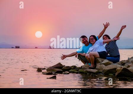 Asiatische Familie Vater Mutter und Teenager-Tochter glücklich sitzen auf Die Uferpromenade am See Wassersonne im orangen Himmel Schöne Naturlandschaft bei Sonnen Stockfoto