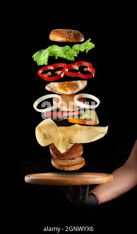 Männliche Hand hält ein rundes Holzbrett und schwimmende Schichten von Cheeseburger mit Hühnereier und Fleischschnitzel auf schwarzem Hintergrund, Fast Food Stockfoto