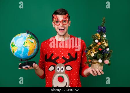 Portrait von attraktiven trendigen fröhlichen Kerl hält kleinen Baum Globus mit Spaß Winter isoliert über grüne Farbe Hintergrund Stockfoto