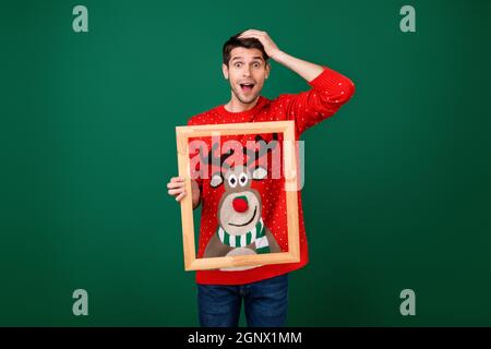 Foto von jungen aufgeregt Mann halten Holzrahmen Bild tragen entworfen Pullover neues Jahr isoliert über grünen Hintergrund Stockfoto