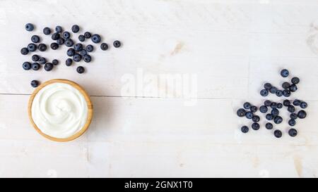 Haufen saftiger Blaubeeren und Holzschüssel aus glattem weißem Joghurt auf weißem Holzküchentisch von direkt oben mit Kopierplatz in der Mitte. Hea Stockfoto