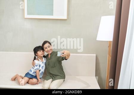 Lächelnde asiatische Senior-Frau, die auf einem modernen Sofa sitzt und mit dem Smartphone fotografiert, während sie mit der Enkelin zusammen fotografiert Stockfoto