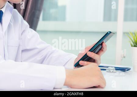 Asiatische Arzt junge gut aussehende Mann lächelnd mit Arbeit oder halten mit digitalen Smartphone im Krankenhaus Schreibtischbüro, Technologie Gesundheitswesen Medica Stockfoto