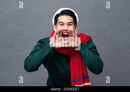 Fröhlicher junger asiatischer Mann in weihnachtlicher Kleidung, schreiend mit Händen, die um den Mund in grauem, isoliertem Studiohintergrund schröpfen Stockfoto