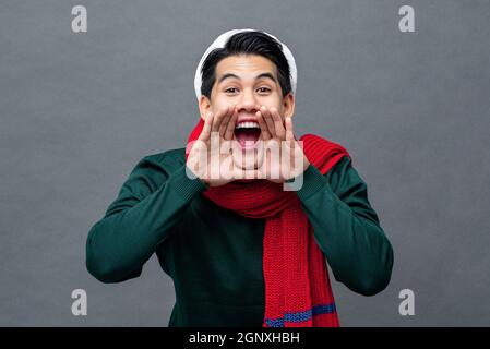 Aufgeregt junger asiatischer Mann in Weihnachtskleidung schreiend mit Händen schröpfen um den Mund in grau isoliert Studio Hintergrund Stockfoto