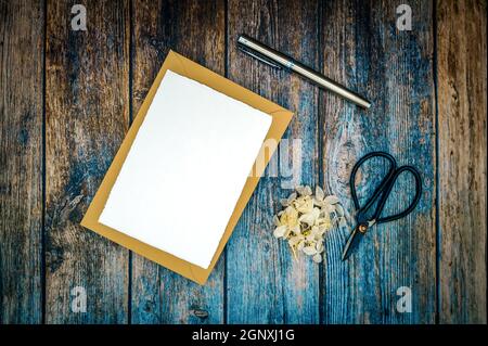 Unbeschriftetes Grußwort, Beileid oder Einladungsmockup mit braunem Umschlag, handgefertigtem Papier und getrockneten Blättern vor einem rustikalen Holztischhintergrund. Stockfoto