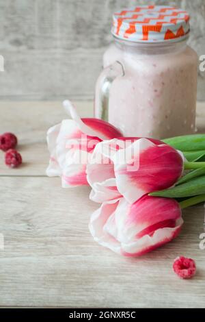 Rosa Himbeer-Smoothie in einem Einmachglas mit frischen Beeren und rosa Tulpen auf einem rustikalen Holzhintergrund, vertikales Bild Stockfoto