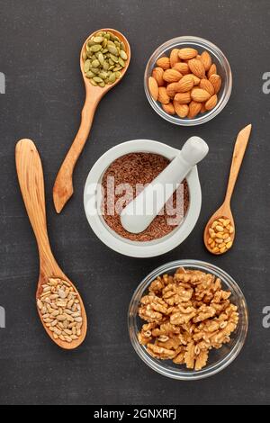 Verschiedene Sorten von Nüssen und Samen liegen in Holzlöffeln und Schalen auf schwarzem Schiefer Hintergrund. Draufsicht. Gesunde Ernährung. Vegetarische Ernährung Stockfoto