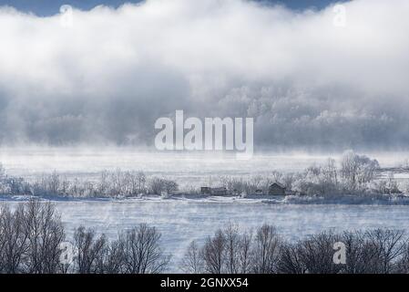 Frostiger Nebel im Winter auf einem nicht gefrorenen Fluss. Bäume und Häuser im Schnee am Ufer. Winterlandschaft Natur. Stockfoto