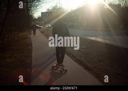 Der Typ auf dem Skateboard in der Sonne. Umweltfreundlicher Transport in der Stadt. Der Mann rollt durch die Stadt. Der Typ fährt auf einem gefährlichen Longboard Stockfoto