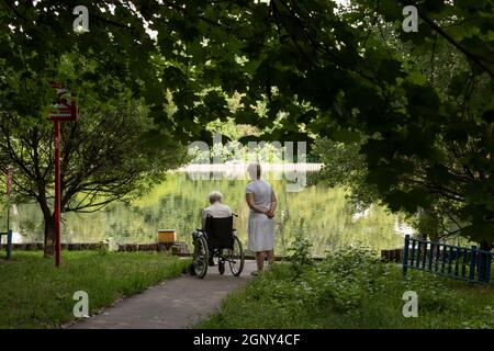 Gehen Sie in der Nähe des Teiches. Ein Mann im Rollstuhl mit einer Krankenschwester geht im Park spazieren. Im Sommer sind die Menschen draußen. Stockfoto