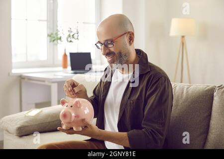 Glücklicher erwachsener Mann, der Geld spart, auf dem Sofa sitzt und Münzen in Sparschwein legt Stockfoto