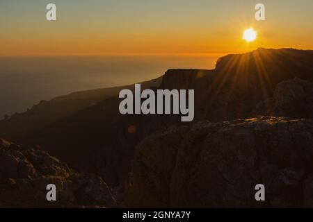 Sonnenuntergang Sonne Berge Strahlen. Hinter den Bergen erhebt sich eine runde orangefarbene Sonne mit schönen Strahlen. Majestätische Berglandschaft im Hintergrund. Stockfoto