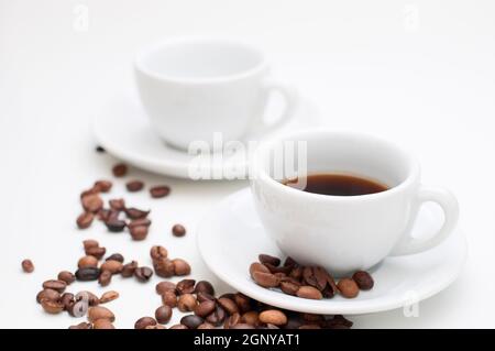 Zwei weiße Cappuccino-Tassen mit Untertassen und verstreuten gerösteten Kaffeebohnen auf weißem Hintergrund. Der Schwerpunkt liegt auf der vorderen Tasse mit Kaffeegetränk gefüllt. Stockfoto