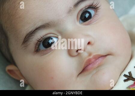 Baby Mädchen mit schwarzen Augen starrte auf die Kamera. Baby mit schönem Gesicht, großen Augen und niedlichen Gesichtsgesten. Stockfoto