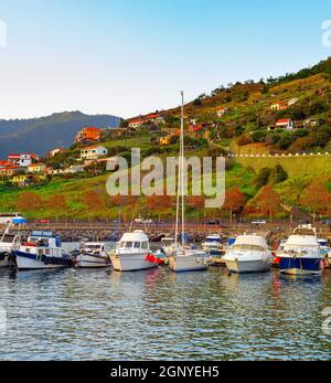 Marina mit Ychten und Motorbooten, die am Ufer des Funcahl festgemacht sind, Sonnenuntergangslandschaft mit Bergen und Dorfhäusern auf dem Hügel, Madeira, Portugal Stockfoto