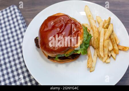 Gut getupfter Hamburger auf einem Brötchen mit Salat, überzogen neben Pommes auf einem weißen Teller neben einer blau-weiß karierten Serviette auf einem Holztisch Stockfoto