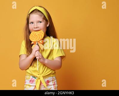 Porträt eines jungen Mädchens mit langen roten Haaren in gelbem T-Shirt und bunten Leggings stehen essen großen Lollipop Stockfoto