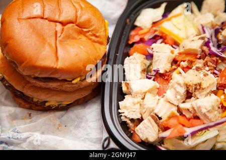 Zwei Cheeseburger gestapelt neben gegrilltem Huhn, Speck, Salat, Kohl, zerkleinerten Käse und gewürfelten Tomatensalat in einem Take-out-Behälter Stockfoto