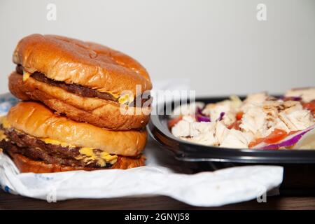 Zwei Cheeseburger gestapelt neben gegrilltem Huhn, Speck, Salat, Kohl, zerkleinerten Käse und gewürfelten Tomatensalat in einem Take-out-Behälter Stockfoto