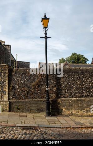 Ein alter, verfärbter, großer schwarzer Laternenpfosten neben einer alten Feuersteinwand in einer kopfsteingepflasterten Straße. Stockfoto