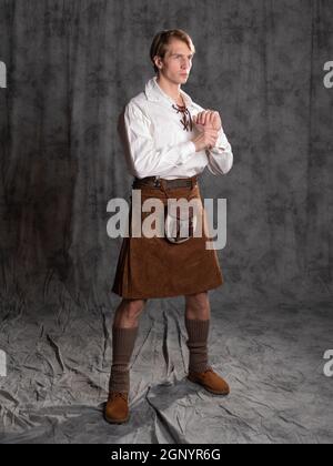 Ein junger Mann in einem Lederkilt und einer weißen Schnürbluse. Ein schottisches Foto in voller Länge im Studio auf grauem Hintergrund Stockfoto