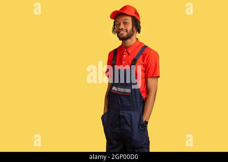 Selbstbewusster, zufriedener Handwerker, der die Kamera anschaut, Uniform trägt, stolz auf geleistete Arbeit ist, die Hände in den Taschen hält, die Dienstleistungsbranche. Innenaufnahme des Studios isoliert auf gelbem Hintergrund. Stockfoto