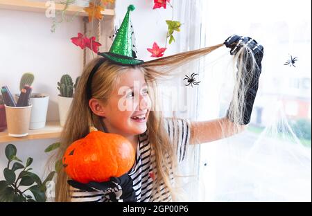 Fröhliches hübsches kaukasisches Mädchen in einem Hexenhut sitzt auf einem für halloween dekorierten Fenster, hält einen Kürbis und sitzt in einem Haargewebe. Vorbereitung Stockfoto
