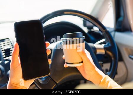 Asiatische Frau trinken heißen Kaffee zum Mitnehmen Tasse in einem Auto Und mit Smartphone leeren Bildschirm, während das Auto in fahren Der Morgen während der Arbeit Stockfoto