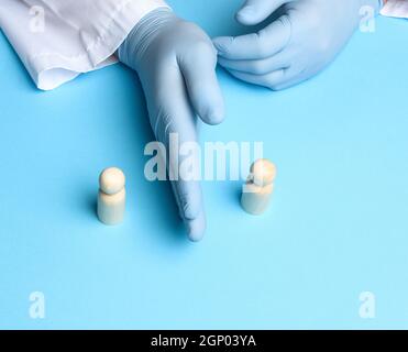 Zwei Holzfiguren von Männern stehen vor dem Hintergrund der männlichen Hände des Arztes in blauen Latexhandschuhen, halten einen Abstand von eineinhalb Metern Stockfoto