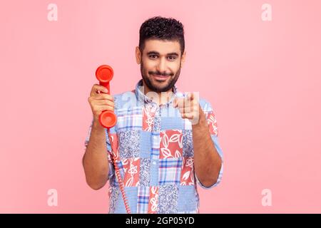 Lächelnder bärtiger Mann in blauem Casual Shirt zeigt mit dem Finger auf Sie, hält in den Händen rotes Retro-Telefon, wartet auf Ihren Anruf, schaut auf die Kamera. Innenaufnahme des Studios isoliert auf rosa Hintergrund. Stockfoto