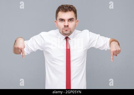 Hier und jetzt. Portrait eines jungen, seriös aussehenden Mannes in weißem Hemd und Krawatte, der mit den Fingern nach unten auf die Kamera schaut. Indoor isoliert auf grauem Hintergrund. Stockfoto
