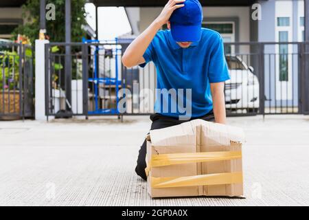 Asiatische junge Lieferung Mann in blauer Uniform er emotional fallen Kurier halten beschädigte Karton ist an der Tür vor Hause gebrochen, Unfall schlechten Transport Stockfoto