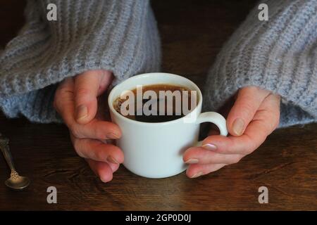 Die Hände der Frau im Pullover halten eine Tasse starken Kaffee auf dem Holztisch. Kaffee-Ventilator Draufsicht Hintergrund. Stockfoto