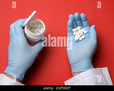 Männliche Hand in einem blauen sterilen Gummihandschuh hält einen Stapel Pillen auf einem roten Hintergrund, Draufsicht Stockfoto
