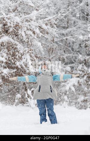Fröhliche junge Mutter in einem Winteranzug mit ihrem Baby in einem Tragetuch auf der Brust und genießt das Leben mit offenen Armen an einem verschneiten Wintertag. Stockfoto