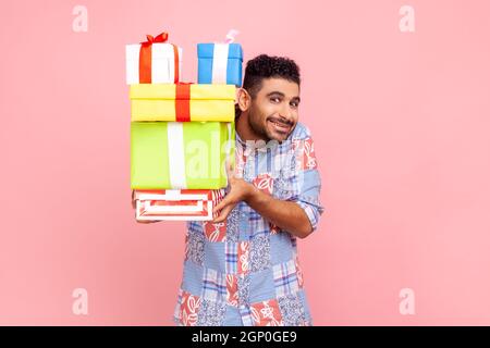 Porträt eines positiven Mannes mit einem toothy Lächeln, das Geschenke aushält, die Kamera anschaut, Geschenke gibt, ein blaues Hemd im lässigen Stil trägt. Innenaufnahme des Studios isoliert auf rosa Hintergrund. Stockfoto