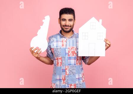 Porträt eines zufriedenen bärtigen Mannes in blauem Casual Shirt mit Papierhaus und großem Schlüssel, Immobilienkauf, Mietdienstleistungen, Hypothek. Innenaufnahme des Studios isoliert auf rosa Hintergrund. Stockfoto