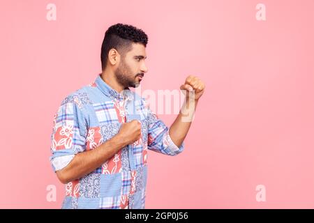 Profilportrait eines aggressiven jungen erwachsenen Mannes in blauem Hemd, der mit Boxfäusten steht und bereit ist, anzugreifen oder zu verteidigen, und mit einem wütenden Gesicht aussieht. Innenaufnahme des Studios isoliert auf rosa Hintergrund. Stockfoto