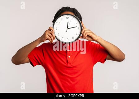 Unbekannter Mann mit Dreadlocks, die ein rotes T-Shirt im lässigen Stil tragen, sich hinter einer großen Wanduhr verstecken, Zeitmanagement, erinnert an Deadline. Innenaufnahme des Studios isoliert auf grauem Hintergrund. Stockfoto