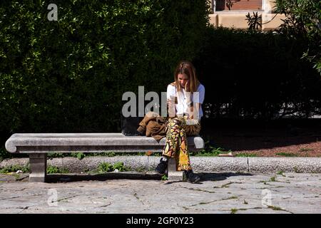 Triest, Italien - Mai 31: Eine junge Frau, die auf der Bank sitzt, sieht sich am 31. Mai 2021 ein Video am Telefon mit Hörgeräten an Stockfoto