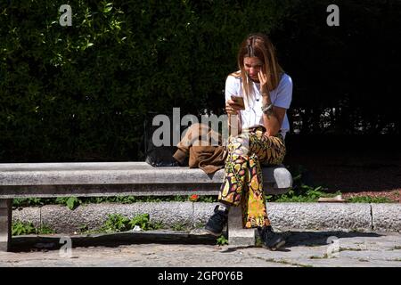 Triest, Italien - Mai 31: Eine junge Frau, die auf der Bank sitzt, sieht sich am 31. Mai 2021 ein Video am Telefon mit Hörgeräten an Stockfoto