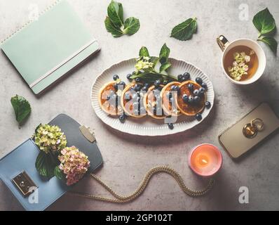 Heidelbeeren Pfannkuchen Frühstück auf dem Schreibtisch des Heims mit einer Tasse Tee, Handtasche, Blumen, Smartphone, Duftkerze, Und Hortensien Blumen. Draufsicht Stockfoto