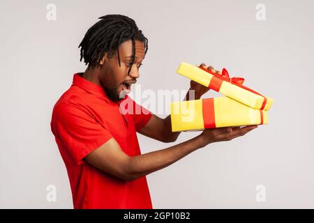 Stauned Mann mit Dreadlocks tragen rote Casual-Stil T-Shirt, öffnen Geschenk-Box und guckt nach innen mit aufgeregt Gesichtsausdruck, zufrieden mit Gegenwart. Innenaufnahme des Studios isoliert auf grauem Hintergrund. Stockfoto