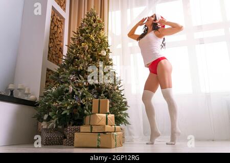 Junge Frau in weißem T-Shirt, Mütze des Weihnachtsmanns und roten Shorts, die auf dem Hintergrund des Fensters neben einem verzierten Weihnachtsfest gähnend und sich ausstrecken Stockfoto